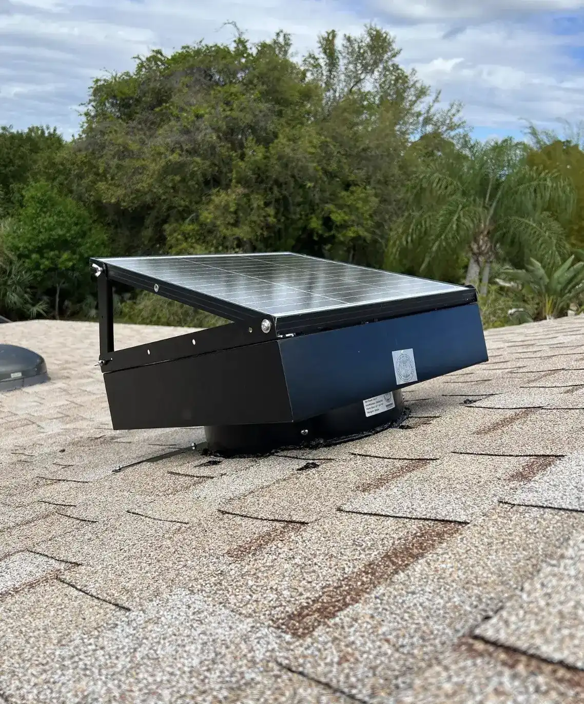 Solar attic fan on a shingle roof with its panel angled toward the sky, pines and palms behind it