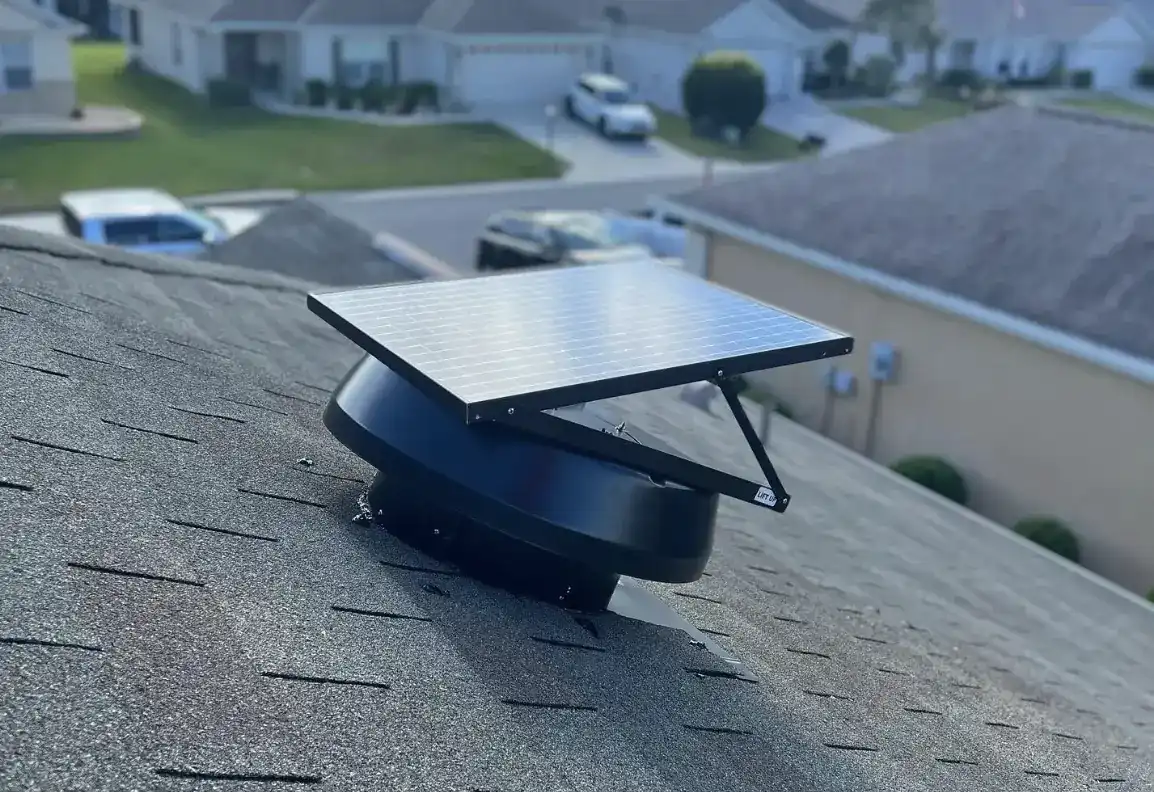 Close view of a solar attic fan, its photovoltaic panel tilted above a round black housing flashed into asphalt shingles