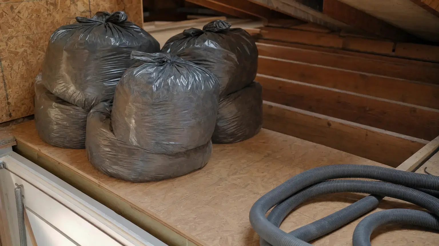Sealed disposal bags of removed attic insulation stacked beside a vacuum hose, with stripped joist bays behind