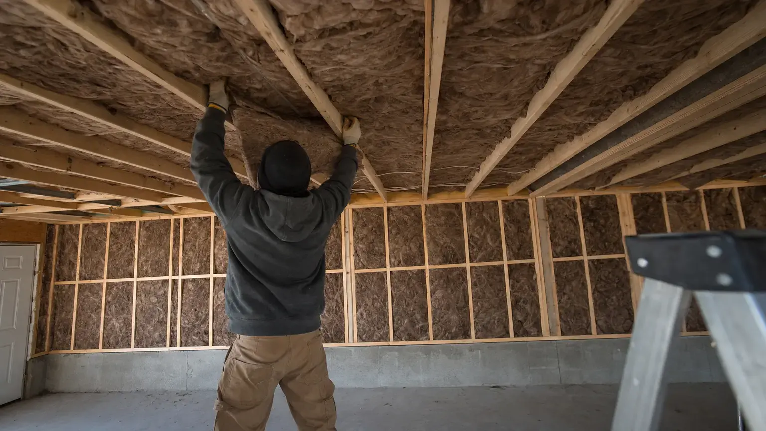Batt insulation being fitted between the overhead joists of a residential garage ceiling, with the wall bays already finished