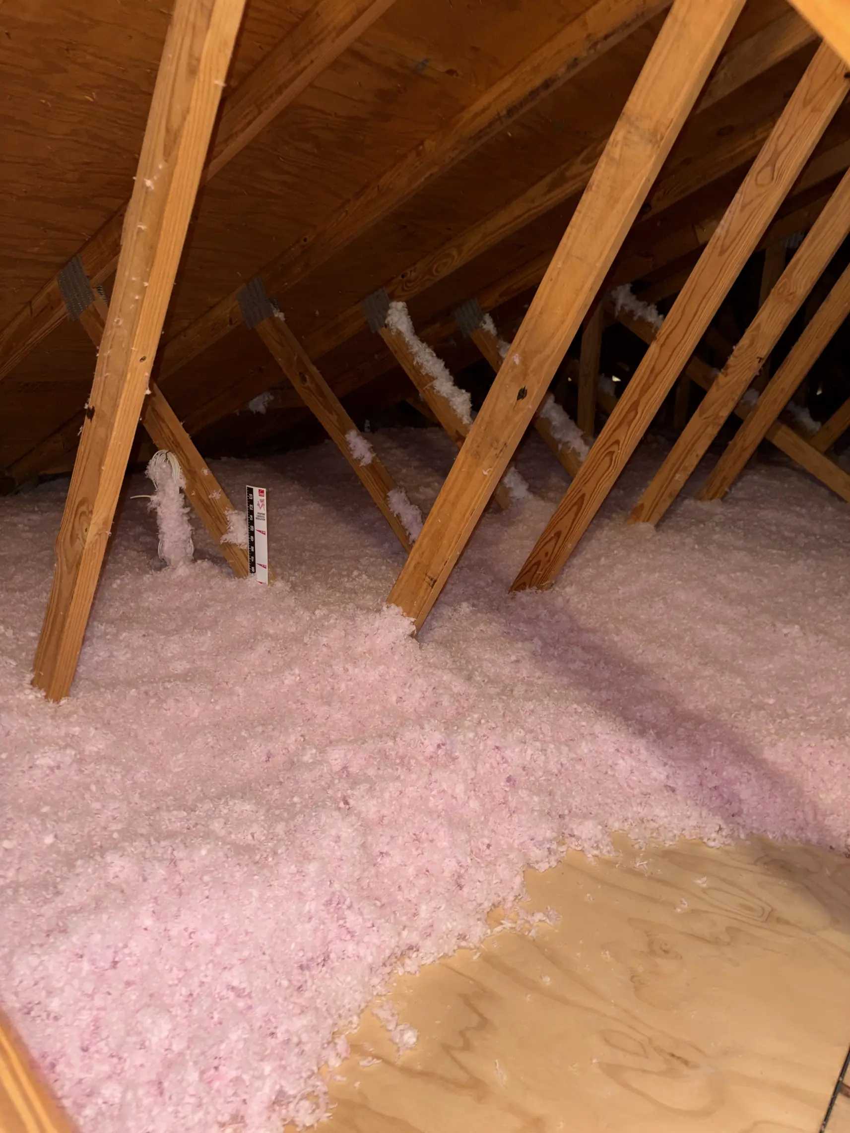 A printed depth ruler standing in blown-in fiberglass insulation, showing the installed thickness against the attic decking